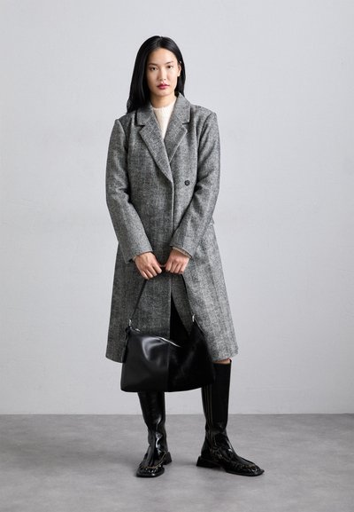 Sivý štruktúrovaný dlhý kabát s vykrojeným golierom, čierna ramenná taška a čižmy po kolená v čiernej farbe. Model stojí pred jednoliatym pozadím.
