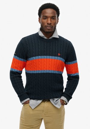 Marineblauwe cable-knit trui met horizontale strepen in oranje en lichtblauw. Inclusief een kraag en een gestreept shirt eronder.