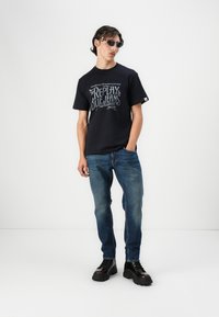 Navy cotton t-shirt with white printed text on chest, paired with blue denim jeans and black lace-up boots. Model wearing sunglasses.