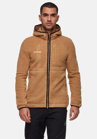 Giacca in pile beige con cappuccio, apertura con zip, tasca sul petto e accenti neri. Presenta una superficie testurizzata e morbida.