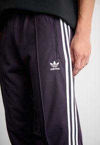 Person som bär svarta Adidas joggingbyxor med vita ränder och logotyp, står med höger hand avslappnad vid sidan.