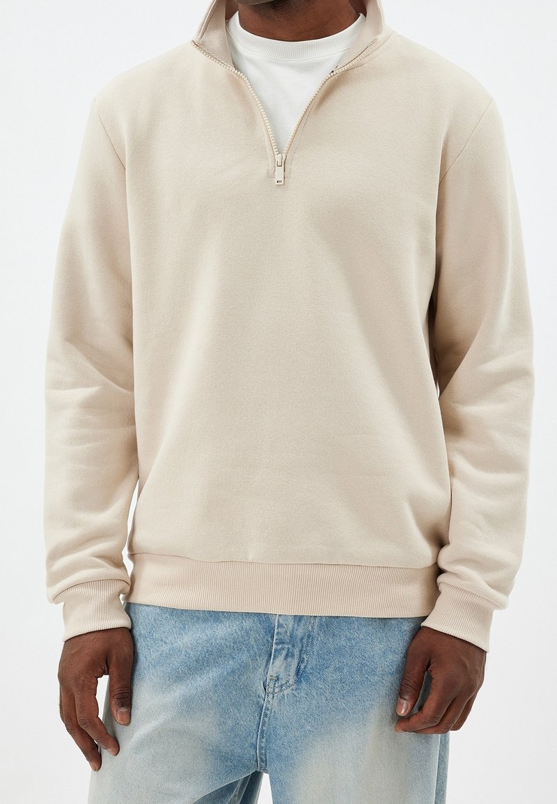 Homme portant un pull beige à fermeture éclair partielle sur une chemise blanche et un jean bleu clair délavé sur un fond uni.