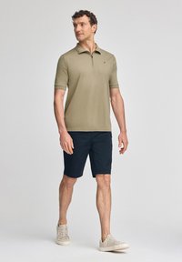 Ljus olivgrön pikétröja med dragkedja, kombinerad med marinblå shorts. Modellen bär beige sneakers och visar en avslappnad stil.
