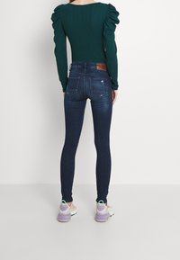 Jeans skinny de color azul oscuro con detalles desgastados, con una cintura alta y dos bolsillos traseros, combinados con una blusa verde ajustada de manga larga.