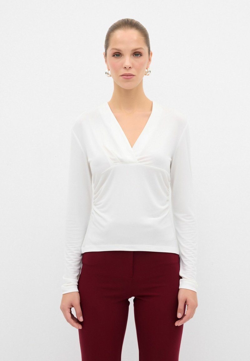Blusa bianca a maniche lunghe con scollo a V; tessuto leggermente texture con arricciature in vita. Abbinata a pantaloni attillati bordeaux.
