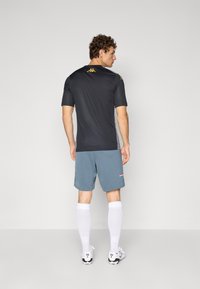 Atletische outfit met een zwart kortemouwen shirt, grijze shorts, witte sokken en zwart-witte schoenen; gouden logo-accenten op de schouders.
