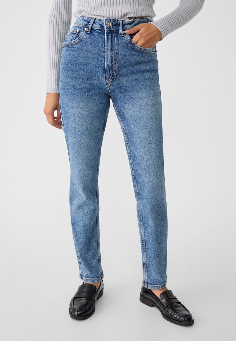 Personne portant un jean slim taille haute bleu clair et un haut gris côtelé à manches longues, avec des mocassins noirs sur fond blanc.