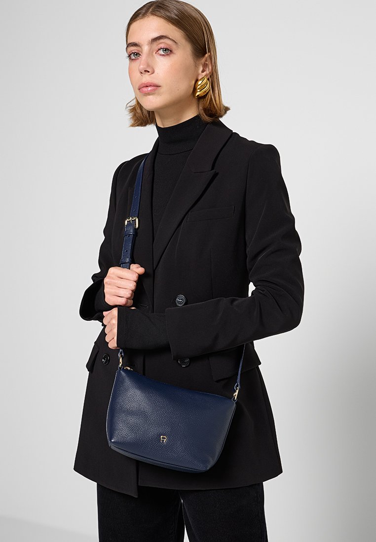 Marineblaue Leder-Umhängetasche mit geschwungener Form, goldenen Beschlägen und strukturierter Oberfläche, getragen von einer Person in einem schwarzen Blazer.