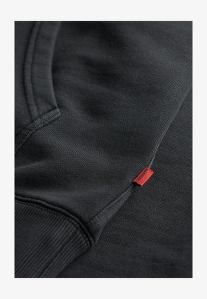 Sudadera negra con un acabado texturizado, que cuenta con una etiqueta roja. El diseño incluye un dobladillo y puños acanalados, enfatizando un estilo casual.