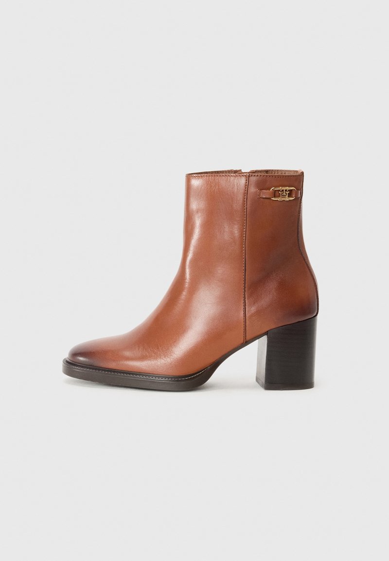 Bottine marron en cuir avec talon bloc et petit détail de boucle dorée près du haut sur un fond clair uni.