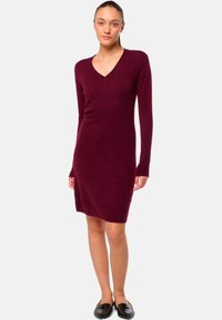 Robe en maille bordeaux avec un décolleté en V et des manches longues, présentant une silhouette ajustée et une texture douce. Portée avec des ballerines noires.