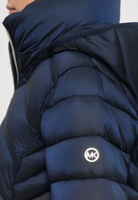 Persoon in een donkerblauwe pufferjack met een hoge kraag en een metallic MK-logo op het bovenste deel van de arm.