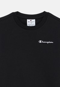 T-shirt in cotone nero con collo a giro, caratterizzato da un logo "Champion" ricamato in bianco sul lato sinistro del petto. Design e texture semplici.