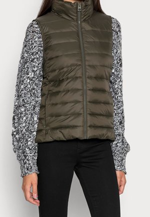 Femme portant une veste matelassée vert olive sur un pull en maille noir et blanc à manches longues et un pantalon noir, debout, bras détendus.
