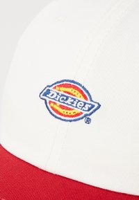 White cap with a red brim, featuring an embroidered circular logo with “Dickies” text in blue and yellow. Cotton material, structured design.