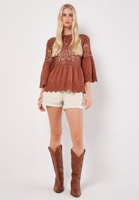 Blouse couleur rouille avec des détails en dentelle complexe, manches évasées et ourlet festonné, associée à un short blanc et des bottes de cow-boy marron.