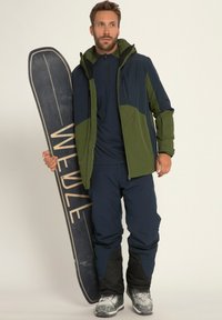 Snowboarder in een marineblauwe en groene jas, bijpassende marineblauwe broek en metalen laarzen, met een donkere snowboard met het "WEDZE" ontwerp.