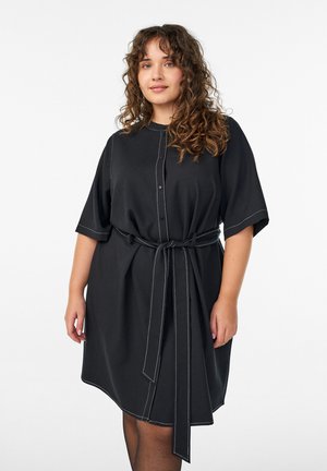 Sort skjortekjole med korte ærmer, knapper foran, bindebånd i taljen og kontrasterende hvide syninger langs sømmene. Glat stoftekstur.