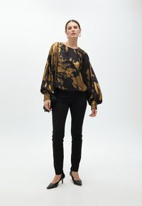 Blusa nera e oro con effetto tie-dye e maniche a palloncino, abbinata a pantaloni neri aderenti e décolleté neri a punta. Accento di una borsa a tracolla semplice.