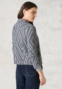 Cardigan en tricot gris avec un motif géométrique en gris clair. Doté d'un col châle et de poignets côtelés, associé à un jean en denim foncé.