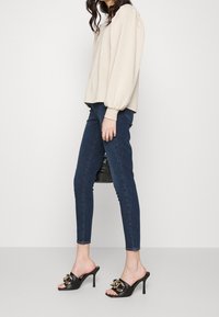 Beige topp med puffärmar, mörka tajta jeans och svarta klackskor med gulddetaljer; modell som står på sidan mot en enkel bakgrund.