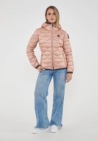 Giovane donna che indossa una giacca trapuntata rosa chiaro, jeans a gamba larga blu con orli sfilacciati e sneakers bianche, in piedi contro uno sfondo semplice.