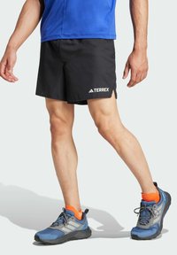 Black athletic shorts with side slits, featuring the "TERREX" logo in white. Worn with blue and gray shoes and bright orange socks.