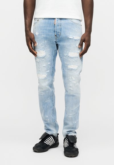 Homme portant un jean déchiré bleu clair et des baskets noires à rayures blanches, debout devant un fond blanc uni.