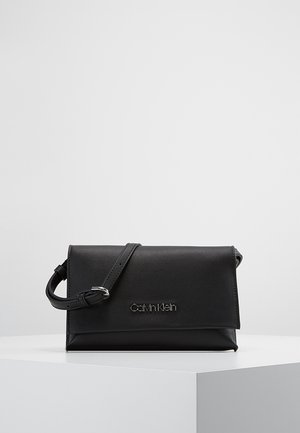 Sac bandoulière en cuir noir Calvin Klein avec fermeture à rabat et sangle réglable, présenté sur un piédestal blanc sur un fond uni.