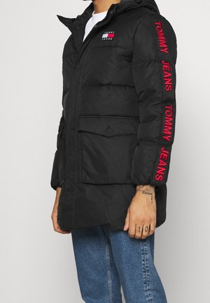 Persoon draagt een zwart gewatteerd jack met het "Tommy Jeans"-logo op de borst en rode tekst op de mouw, gecombineerd met een blauwe spijkerbroek.