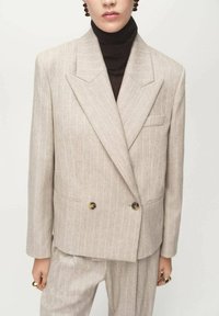 Blazer de doble botonadura en tonos beige con rayas finas, solapas anchas, bolsillos frontales y botones en tono dorado, combinado con un suéter de cuello alto oscuro.