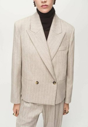 Blazer de doble botonadura en tonos beige con rayas finas, solapas anchas, bolsillos frontales y botones en tono dorado, combinado con un suéter de cuello alto oscuro.