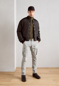 Black bomber jacket with snap buttons, olive green zip-neck shirt, light gray cargo pants with pockets, and black ankle boots.