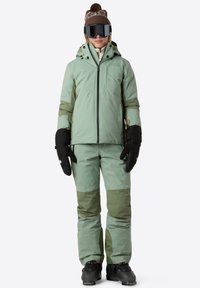 Voděodolný dvoudílný lyžařský outfit ve světle zelené a tmavě zelené barvě, obsahující zipovou bundu, odpovídající kalhoty a černé palčáky.
