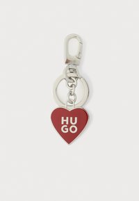 Porte-clés en forme de cœur rouge avec ferrure argentée, arborant le texte "HUGO" en blanc. Comprend un crochet à pression et un anneau de clé circulaire.