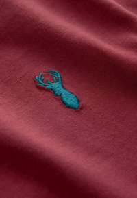 Emblemat turkusowej głowy jelenia wyszywany na teksturowanej bordowej tkaninie, podkreślający szczegółowe ściegi i splot materiału.