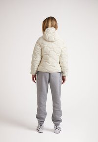 Ljusbeige pufferjacka med ett kviltat mönster och huva, tillsammans med grå sweatpants. Outfiten har elastiska manschetter och sportiga skor.