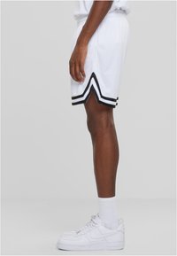Shorts de sport blancs avec des rayures noires, présentant une texture en maille et une fente en forme de V à l'ourlet. Associés à des baskets blanches et des chaussettes.