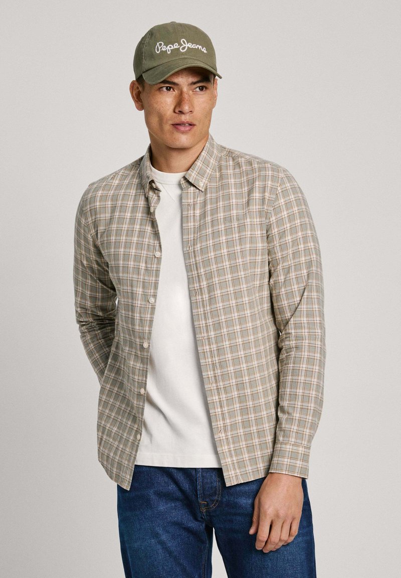 Camicia a quadri in beige chiaro, verde e arancione con una t-shirt bianca a girocollo sotto, abbinata a un berretto color khaki. Jeans di denim.