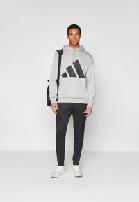 Siva kapuca s kapuco z velikim črnim adidas logotipom, v kombinaciji s črnimi trenirkami z majhnim logotipom ter belimi športnimi čevlji.