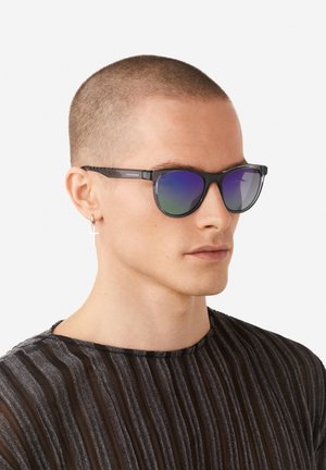 Gafas de sol negras con lentes degradados en azul y verde, que presentan un sutil patrón texturizado en las patillas. Se llevan con una camisa negra a rayas.