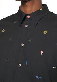 Homme portant une chemise noire boutonnée avec un col et de petites icônes colorées comprenant des cibles, des pièces de puzzle et des ampoules.