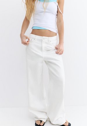 Femme portant un jean blanc ample et un top court superposé blanc et bleu, debout devant un fond blanc uni.