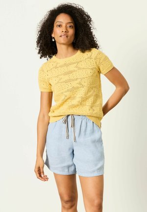 Frau mit lockigem Haar, die ein gelbes kurzärmeliges Stricktop und hellblaue Shorts mit Kordelzug trägt, steht mit einer Hand in der Hüfte.