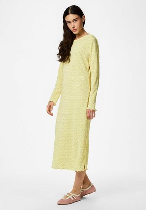 Žena s dlouhými tmavými vlasy, oblečená ve žlutých midi šatech s dlouhým rukávem a růžových plochých botách se stříškami, stojící před bílým pozadím.