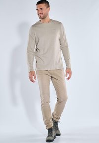 Beige Langarmshirt mit rundem Ausschnitt, kombiniert mit hellbeigen Hosen und dunklen Stiefeln. Glatte Textur, dezente Markenkennzeichnung auf dem Shirt.