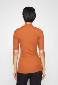 Persona con capelli neri corti che indossa una maglietta arancione a maniche corte e pantaloni neri, in piedi con le spalle rivolte verso un semplice sfondo bianco.