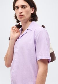 Νεαρός άντρας με καστανά μαλλιά μέχρι τους ώμους φορώντας ένα ανοιχτό μωβ πουκάμισο με κοντά μανίκια και κουμπιά, κουβαλώντας μία μπεζ και καφέ τσάντα στον ώμο του.