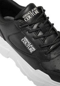 Sneaker en cuir noir avec une semelle en caoutchouc blanc. Caractéristiques incluant un bout arrondi, des lacets noirs et une étiquette indiquant "VERSACE JEANS COUTURE."