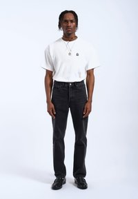 Camiseta de algodón blanca con un pequeño logo negro, combinada con jeans de mezclilla negros y mocasines negros, de pie contra un fondo liso.
